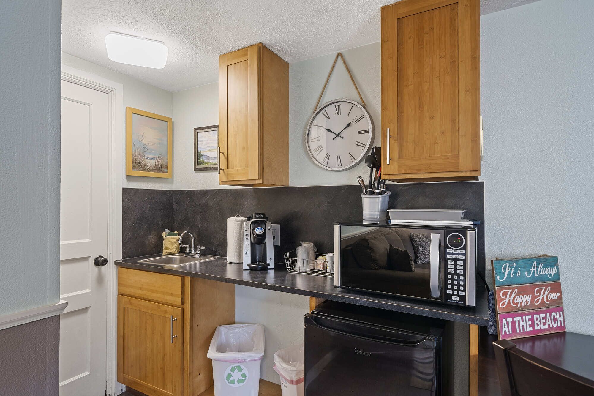 A compact kitchen with wooden cabinets, a wall clock, a microwave, a coffee maker, and a trash bin.