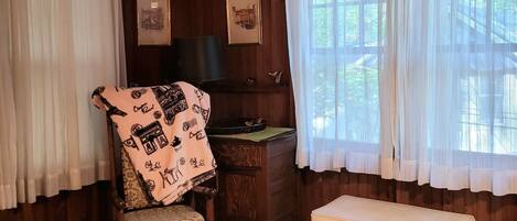 A cozy room with wooden walls, a bed, a desk with a chair, a bench, and a large window with sheer curtains.