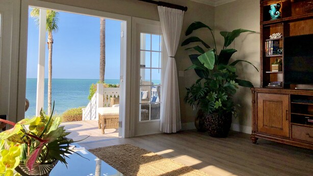 A living room with a large window overlooking the ocean, a television, and a bookshelf. - Waterfront Luxury Villa (62) with sunset views and boat slip (Marathon)
