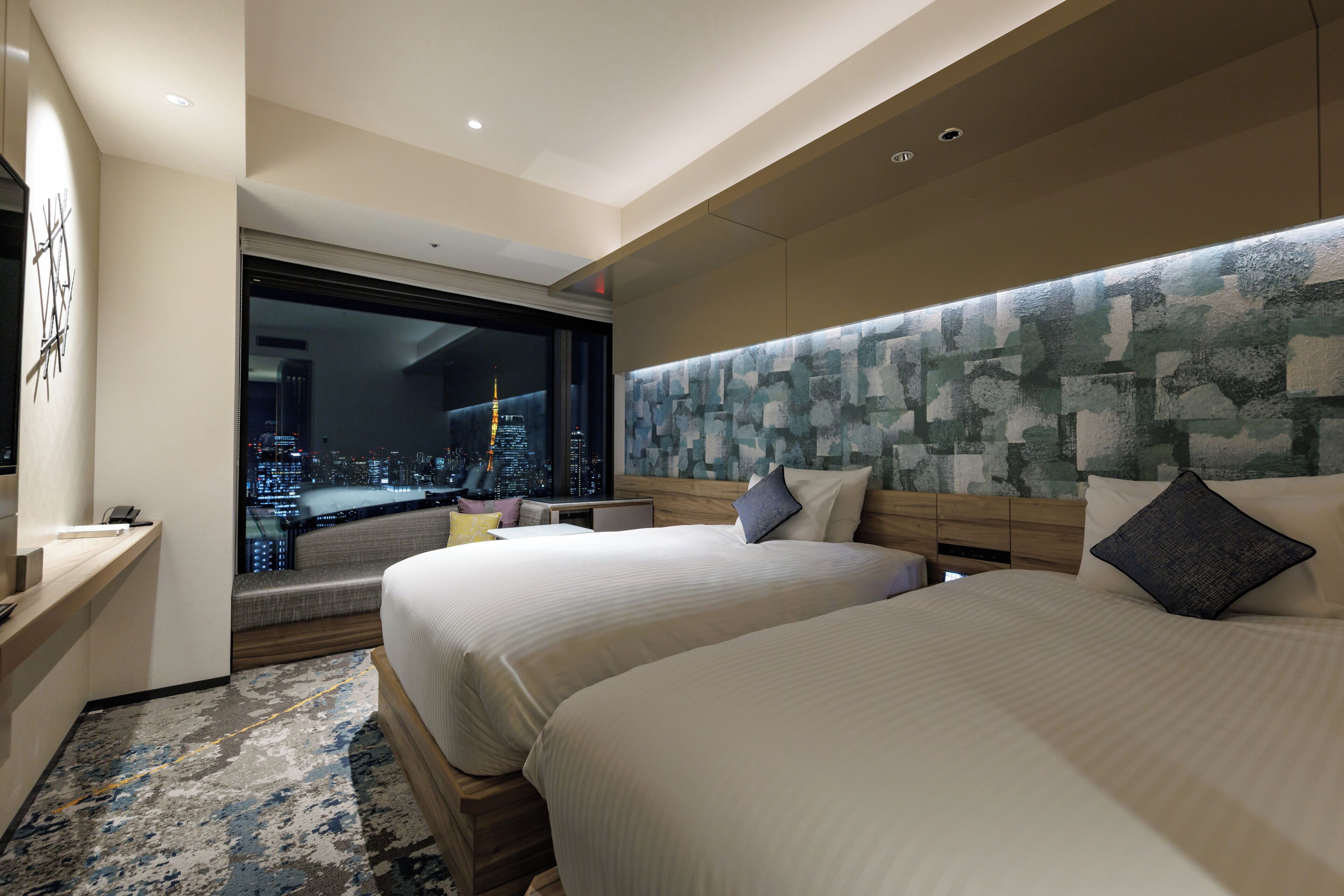 A hotel room with two beds, a large window with a city view, a wooden headboard, and a small desk with a telephone.