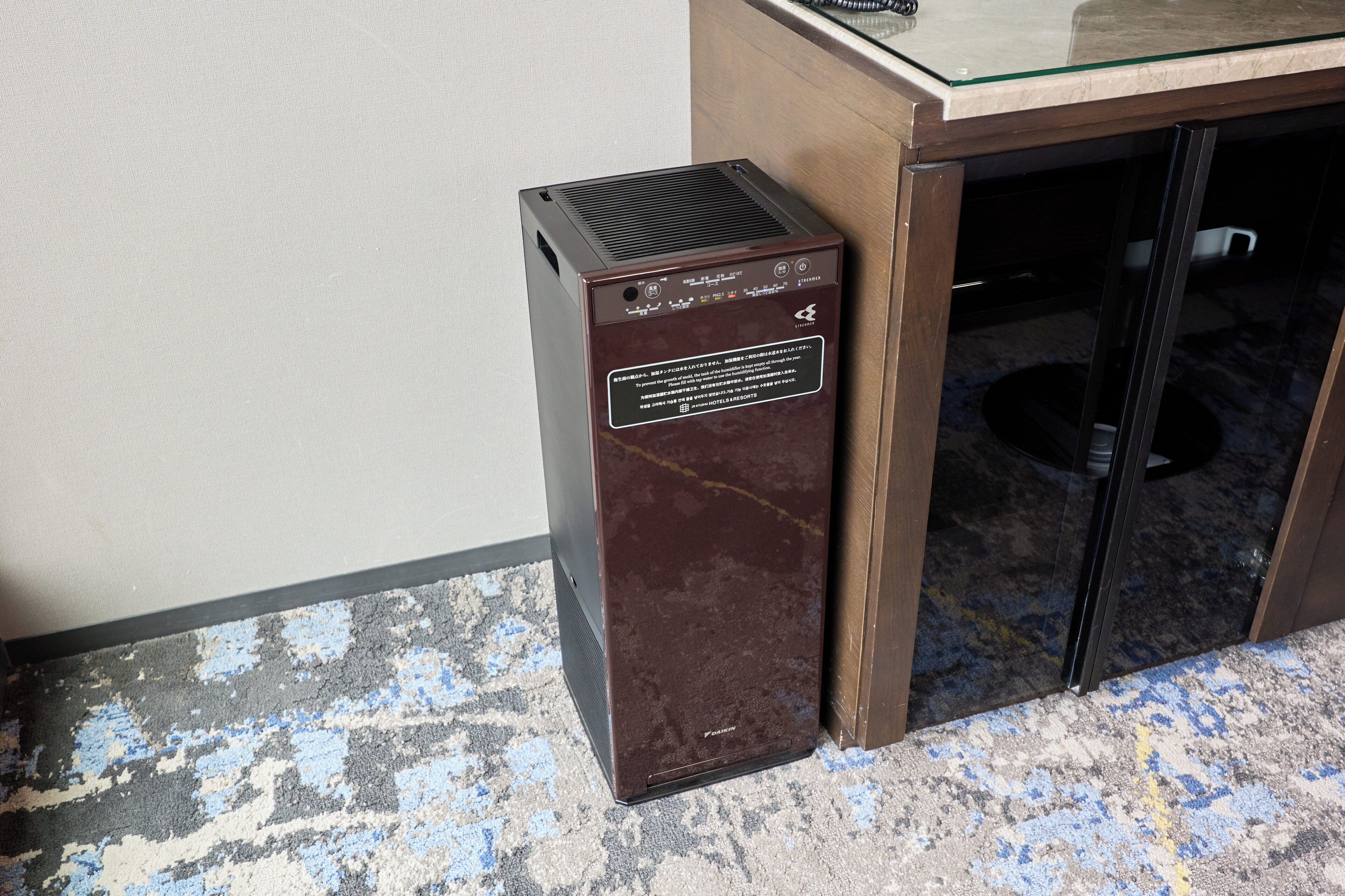 A compact, dark-colored heater unit with a control panel and a label, placed on a patterned floor next to a glass door.