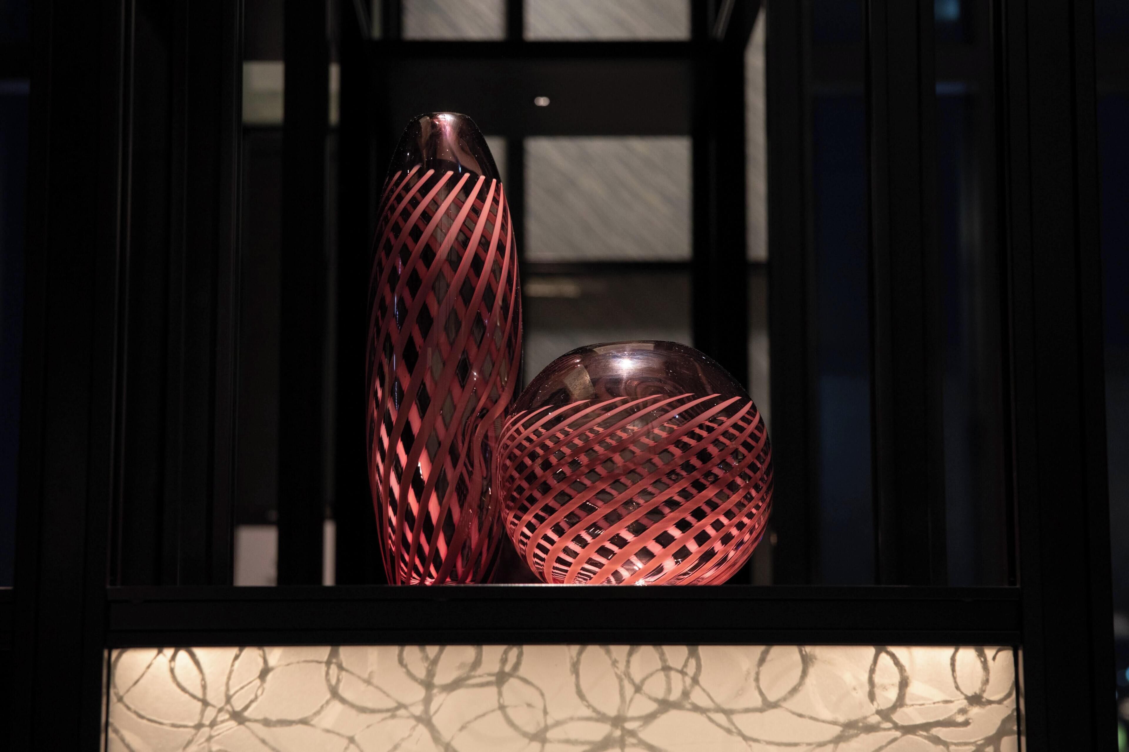 Dos jarrones de vidrio con un diseño de celosía, exhibidos en un espacio interior oscuro con una superficie reflectante debajo.