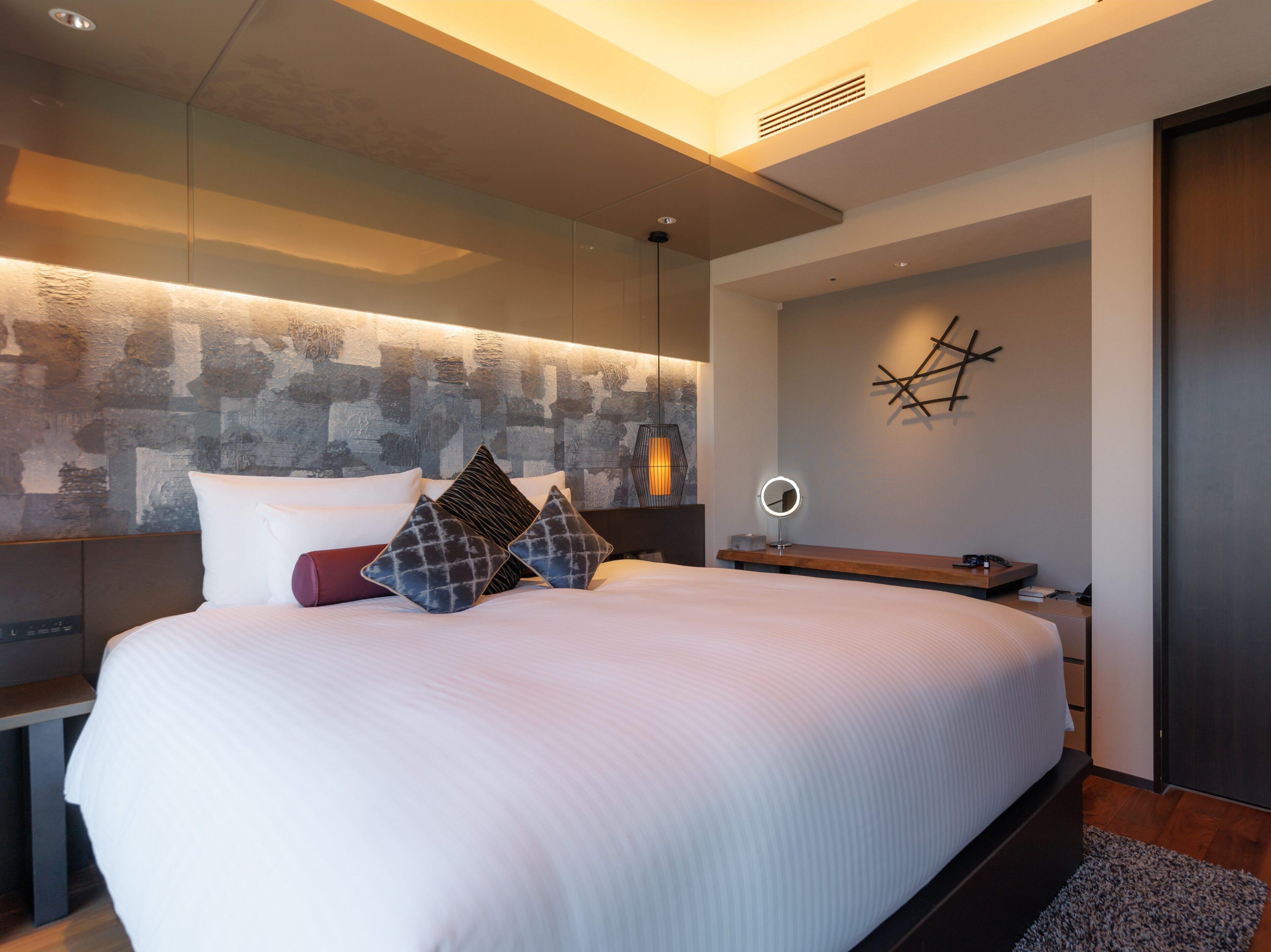 Une chambre à coucher moderne avec un grand lit, une table de chevet, une lampe fixée au mur et un élément décoratif mural.