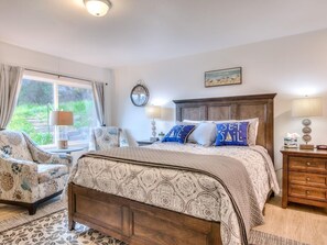 A bedroom with a large bed, a nightstand, a chair, a lamp, and a view of greenery outside. - W7 - Minke - Whale Inn (Depoe Bay)