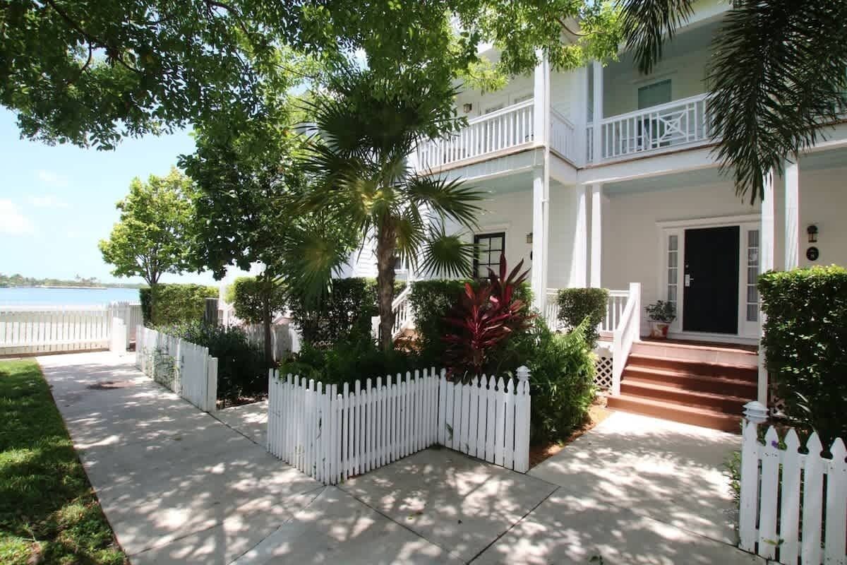 A two-story white house with a balcony, a white picket fence, and a well-maintained garden with various plants.