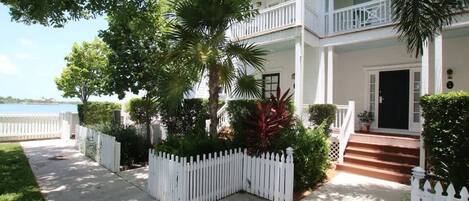 A two-story white house with a balcony, a white picket fence, and a well-maintained garden with various plants.