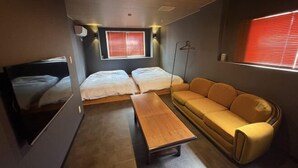 A compact room with two beds, a sofa, a table, and a window with blinds.