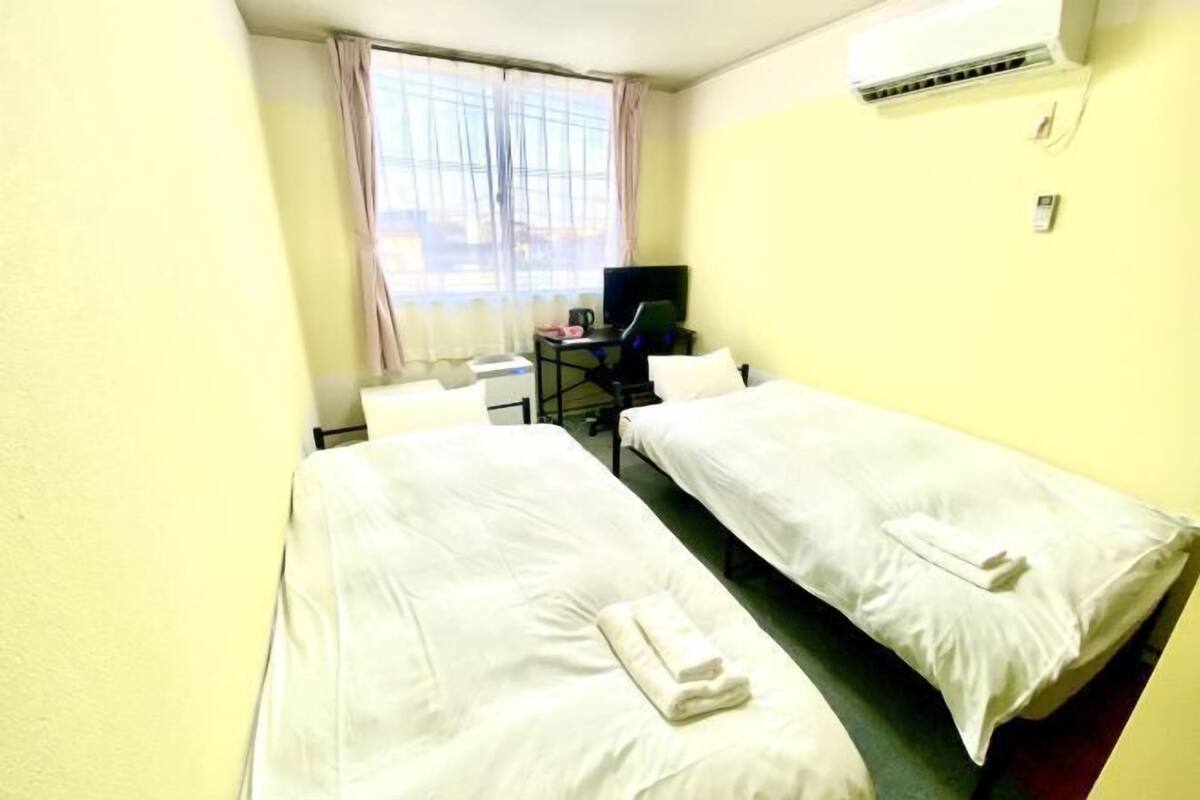 A hotel room with two single beds, a desk with a chair, a television, and an air conditioning unit.