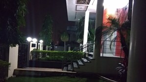 A nighttime view of a building with a staircase leading to a gated entrance, surrounded by greenery and palm trees.