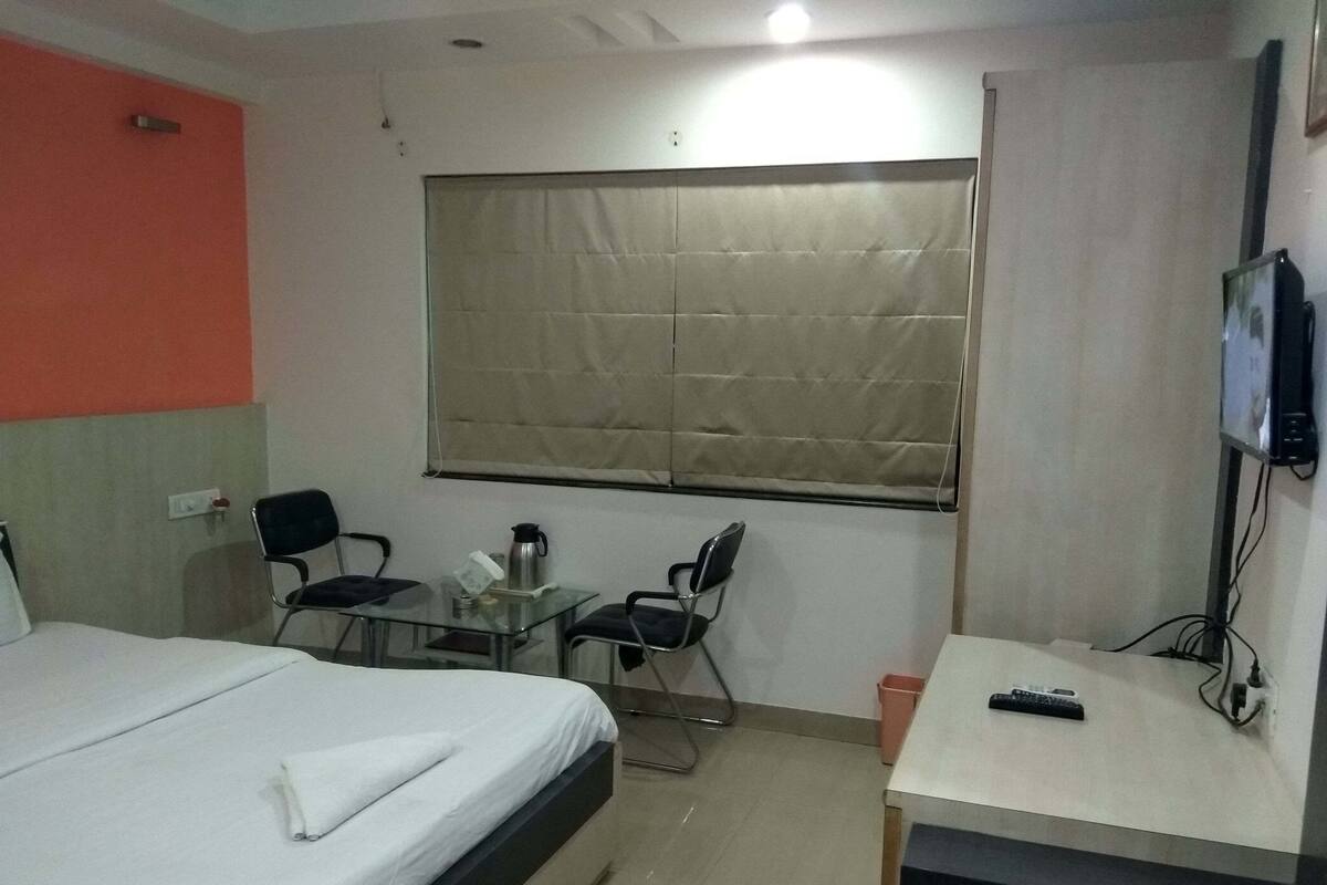 Quarto de hotel com uma cama, uma pequena mesa com cadeiras, uma televisão e uma janela com persiana bege.