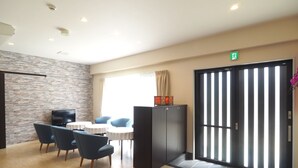 Eine Hotelhalle mit Steinwand, einer Rezeption, einer Sitzecke mit Stühlen und Tischen sowie einer großen Schiebetür mit vertikalen Jalousien.