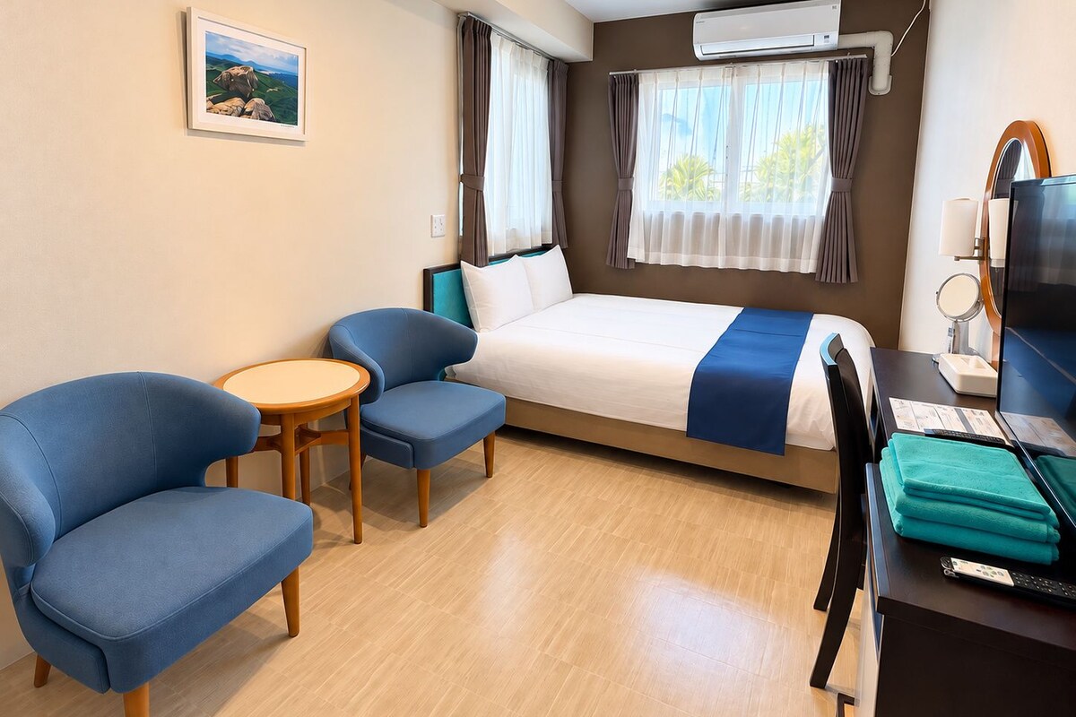 Une chambre d’hôtel avec un lit, deux fauteuils bleus, une petite table, un bureau avec un ordinateur et un appareil de climatisation.