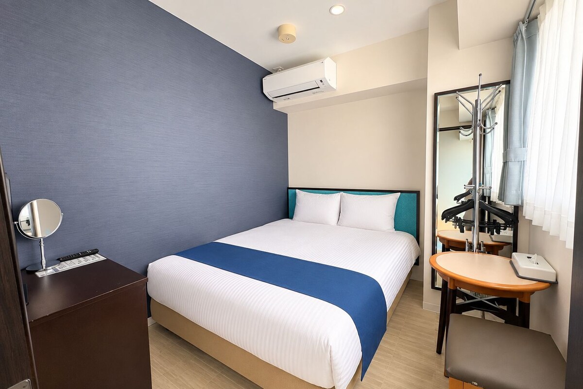 Une chambre d’hôtel comprenant un lit, une table de chevet avec un miroir, une petite table avec une chaise, un appareil de climatisation et un porte-manteau.