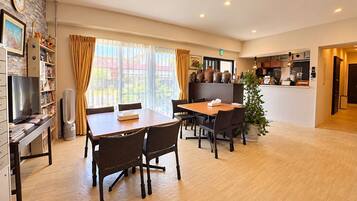 A hotel room with a dining area, a TV, a bookshelf, and a large window with curtains.