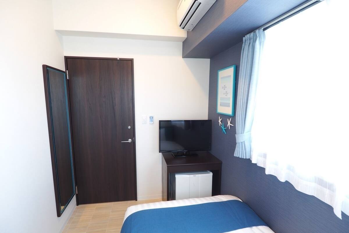 Een slaapkamer met een bed, een televisie, een klein bureau met een computer, een deur en een raam met gordijnen.