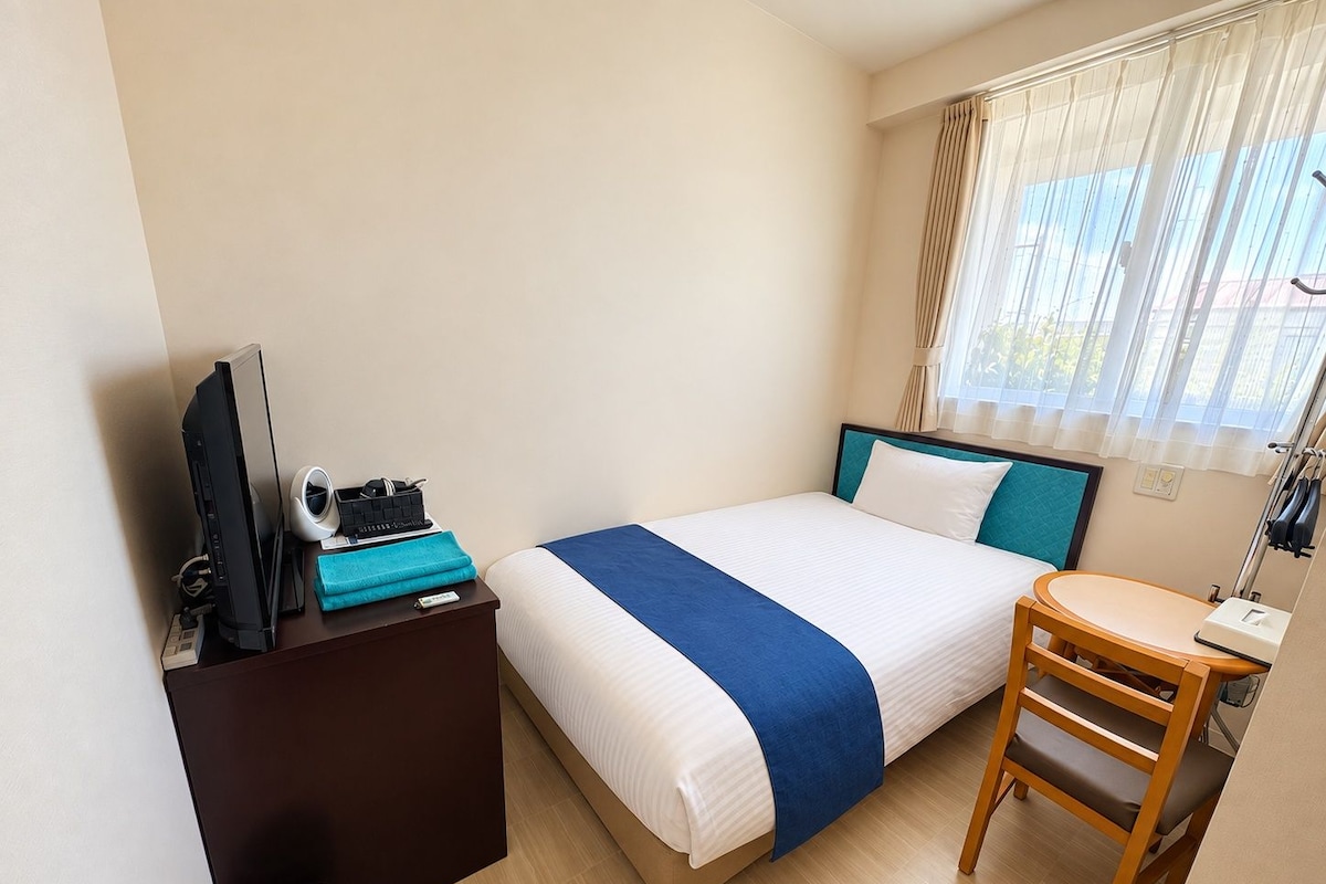 Ein Hotelzimmer mit einem Bett, einem Schreibtisch, einem Stuhl, einem Fernseher und einem Fenster mit Vorhängen.