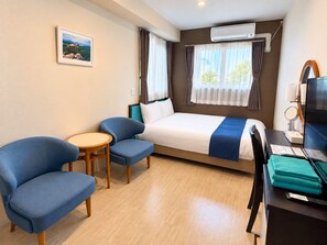 Une chambre d’hôtel avec un lit, deux fauteuils bleus, une petite table, un bureau avec un ordinateur et un appareil de climatisation.