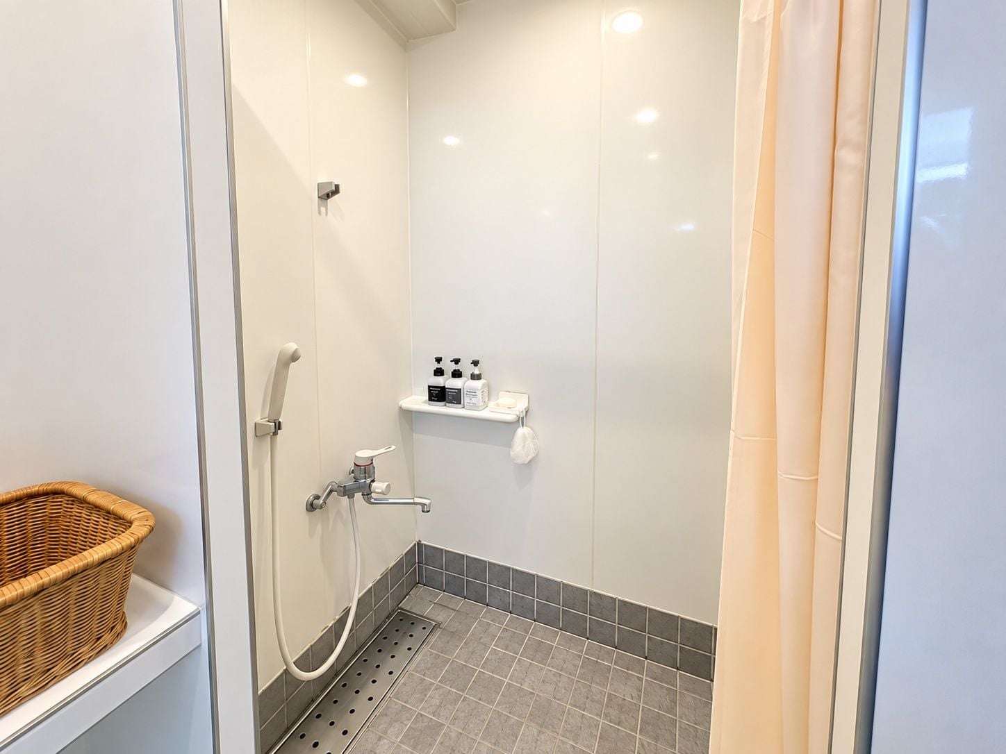 A small, modern bathroom with a shower, a white shelf with toiletries, and a wicker basket on a white counter.