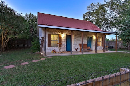 A stone house with a red roof, a covered porch, and a fenced backyard.
