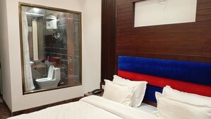 A hotel room with a large window, a bed with white bedding, a wooden headboard with blue and red panels, and a bedside table with a phone. - JJK Rukmini Vilas Hotels and Banquets (Muzaffarpur)
