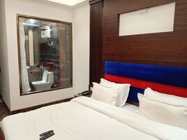 A hotel room with a large window, a bed with white bedding, a wooden headboard with blue and red panels, and a bedside table with a phone.