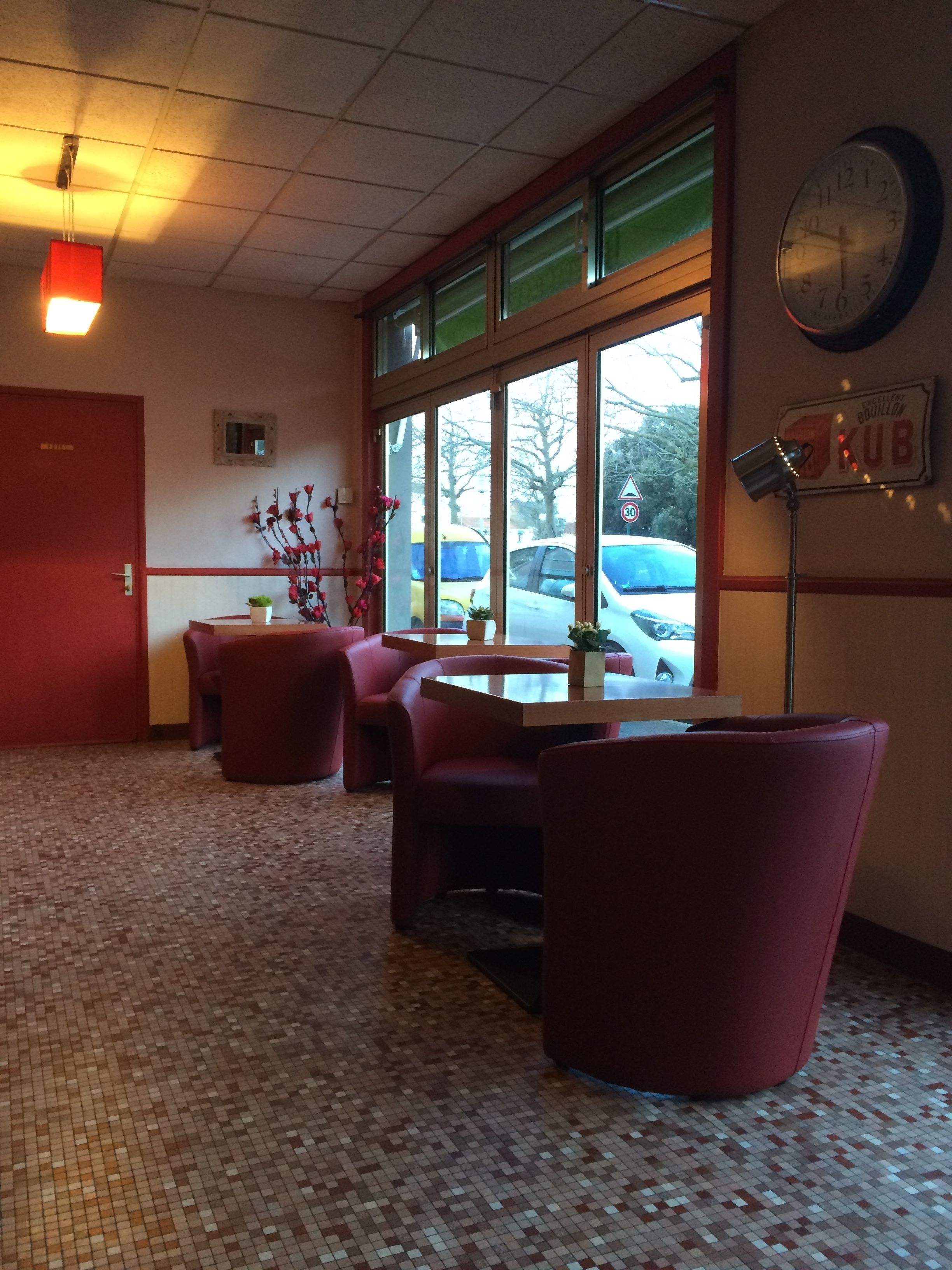 Une pièce avec un sol carrelé, des chaises rouges, une table, une horloge et une fenêtre donnant sur les voitures garées à l’extérieur.