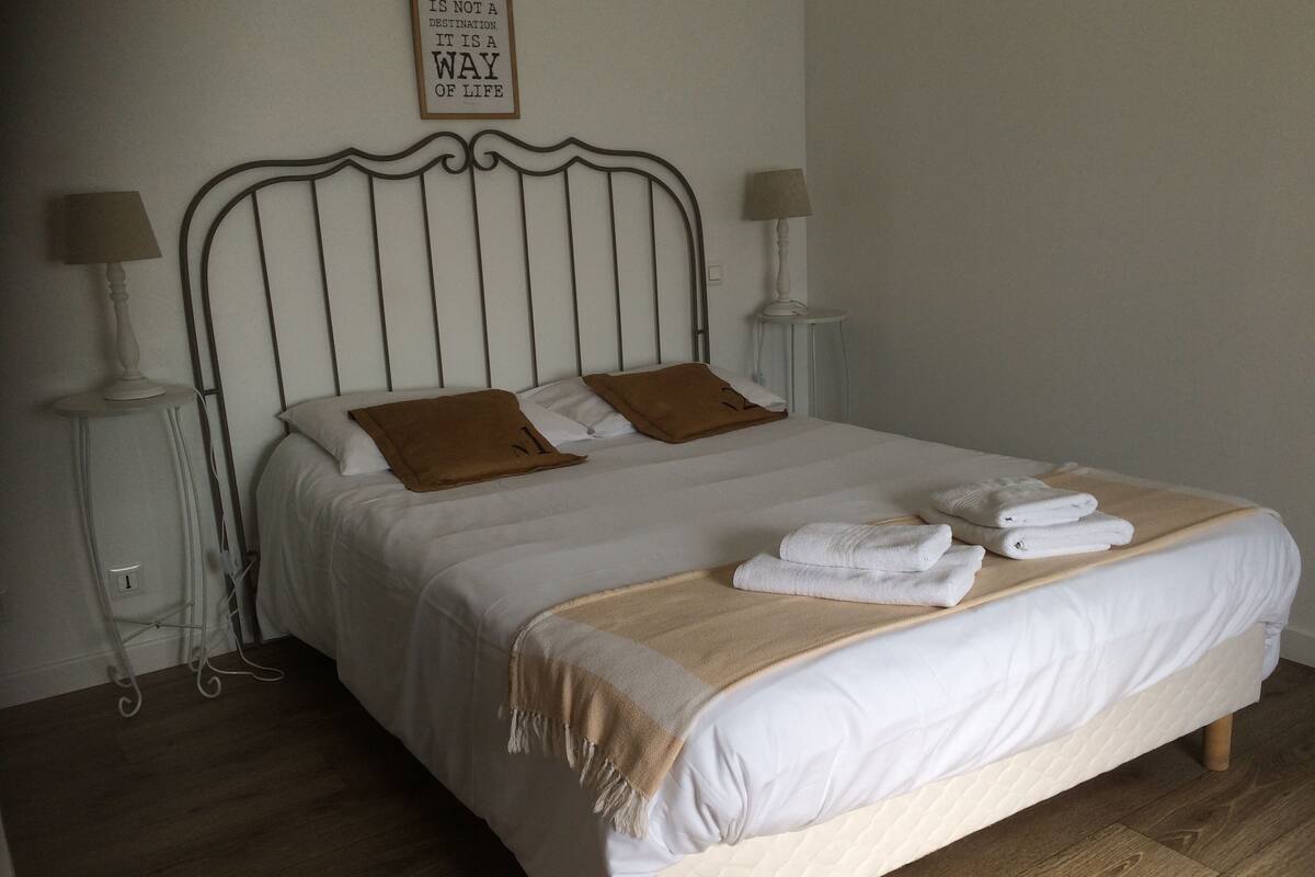 Una cama bien hecha con sábanas blancas, dos almohadas marrones y una manta color beige. Hay dos lámparas de noche a cada lado de la cama.