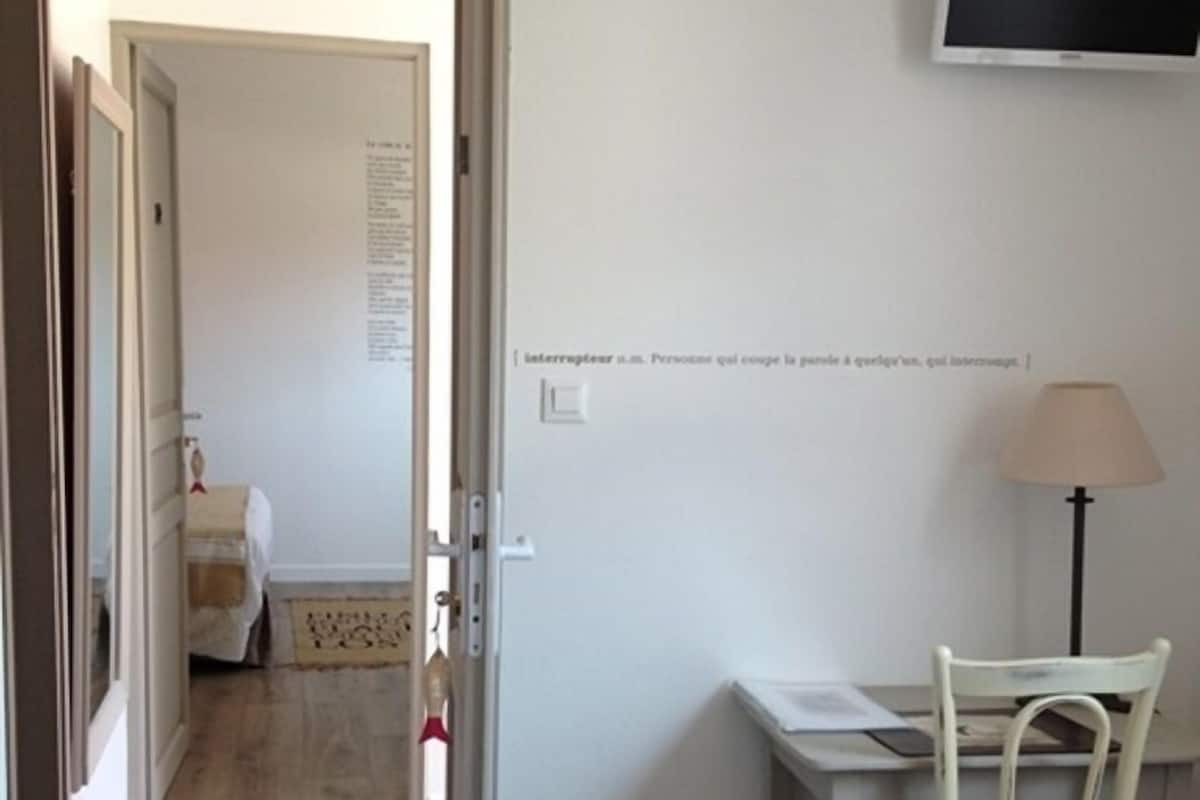 Una habitación con piso de madera, un escritorio con una silla, un televisor en la pared y una puerta que da a otra habitación.