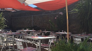 Un patio al aire libre con mesas y sillas, un paraguas grande que da sombra y una pared de piedra al fondo.