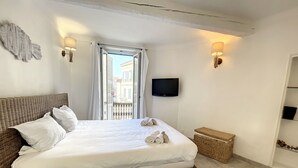 Un letto rifatto con lenzuola bianche, una testiera in vimini e vista sugli edifici attraverso una finestra con tende bianche.