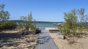 Ein Holzweg führt zu einem Strand mit freiem Blick auf das Wasser und Boote in der Ferne.