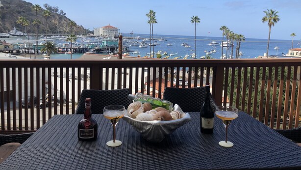 A table with a bottle of Grand Marnier, a glass of wine, and a bowl of seashells, overlooking a marina with boats and palm trees. - Catalina's Premier Bay View Vacation Home (Avalon)