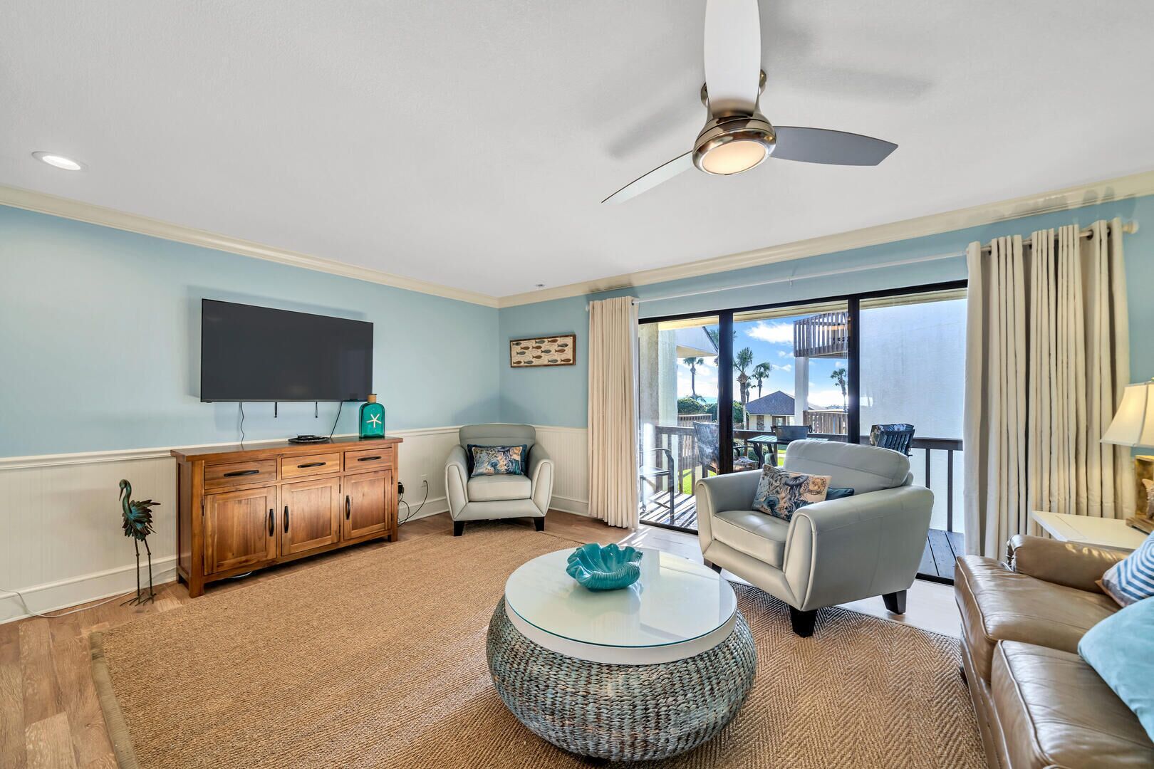 A living room with a flat-screen TV, wooden cabinet, two armchairs, a round coffee table, and a ceiling fan.