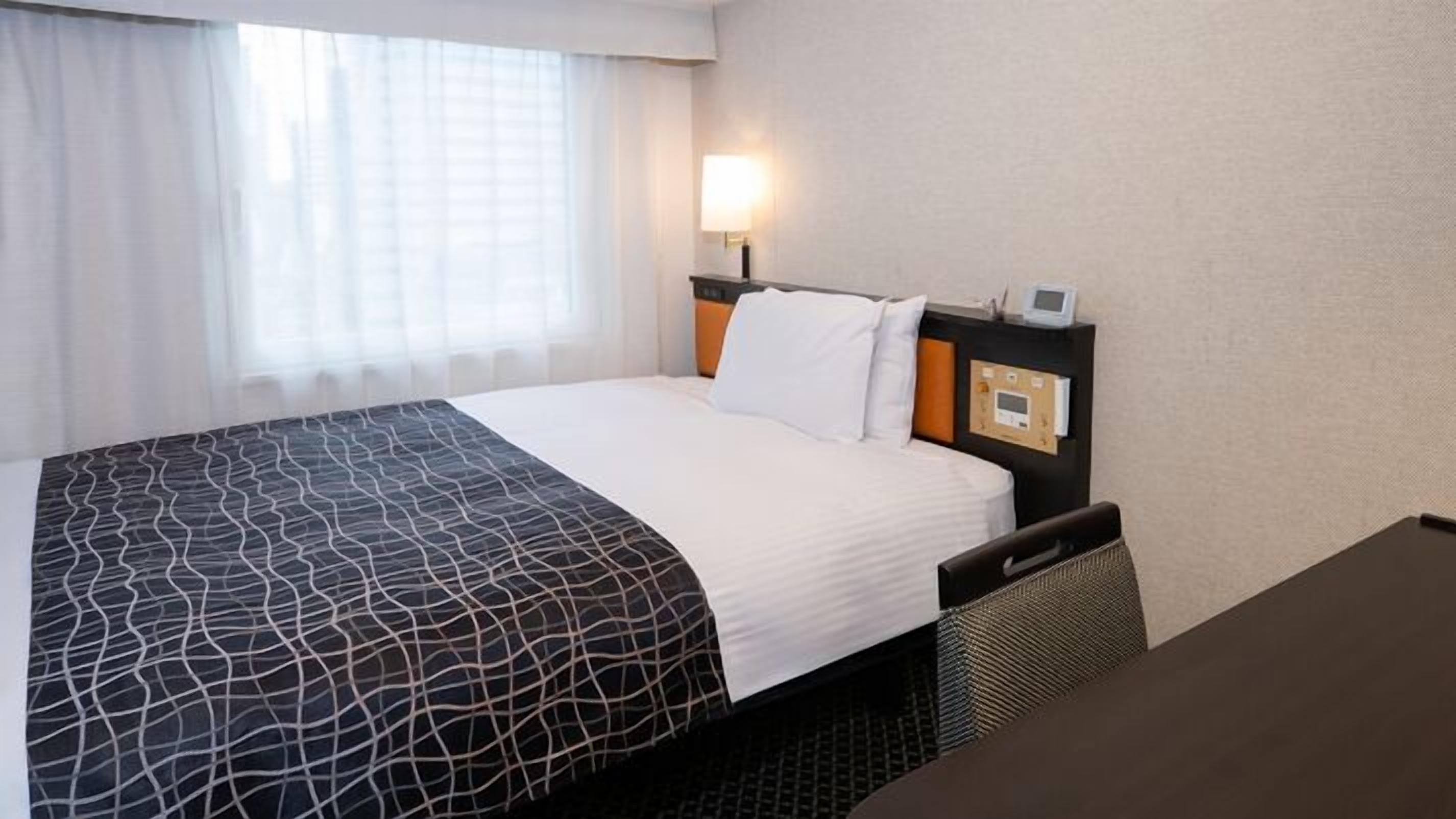 Ein Hotelzimmer mit Bett, Nachttisch, Lampe und einem fenster mit Vorhängen.