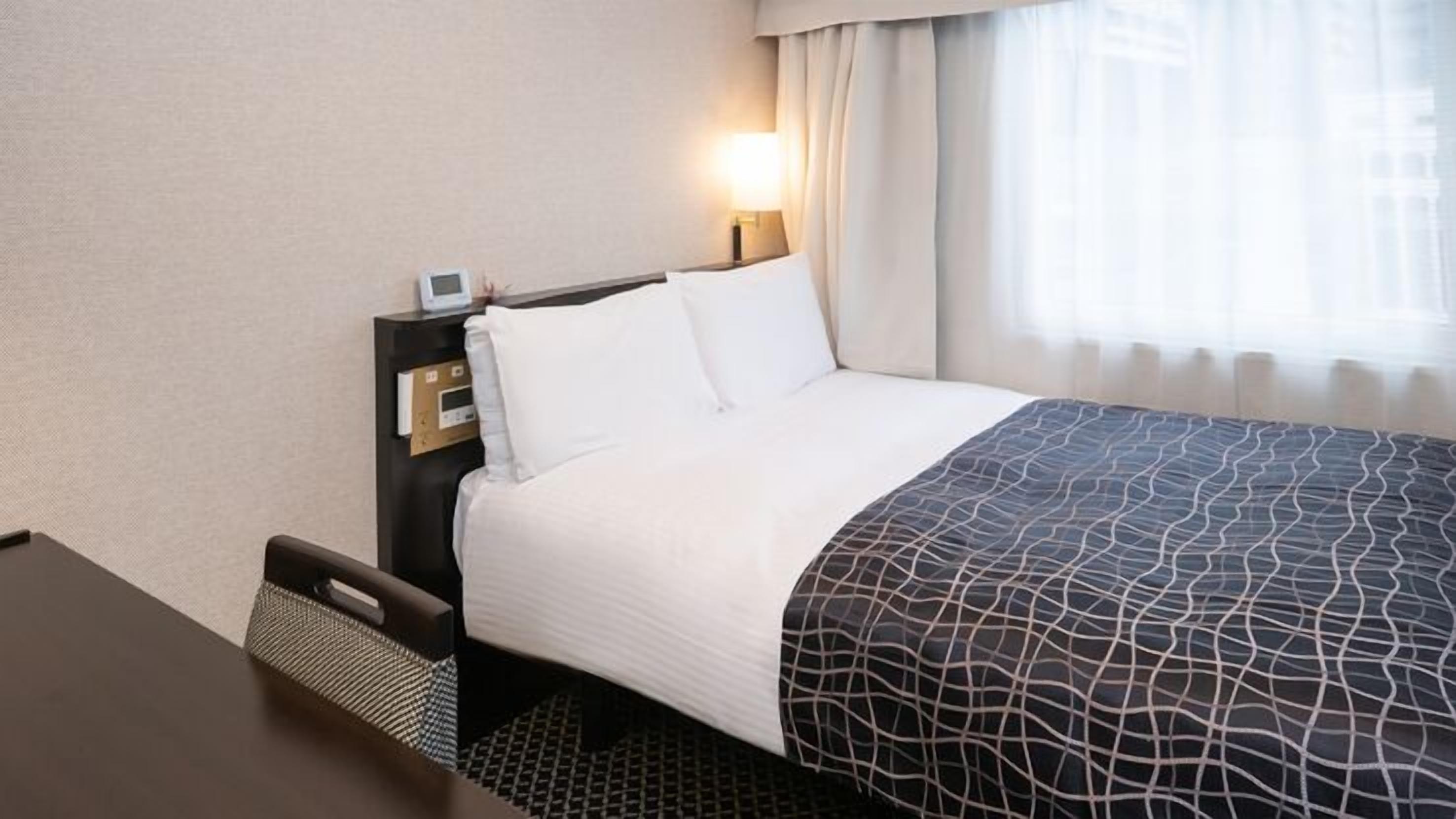 Ein Hotelzimmer mit Bett, Schreibtisch, Nachttisch mit Uhr und Fenster mit Vorhängen.