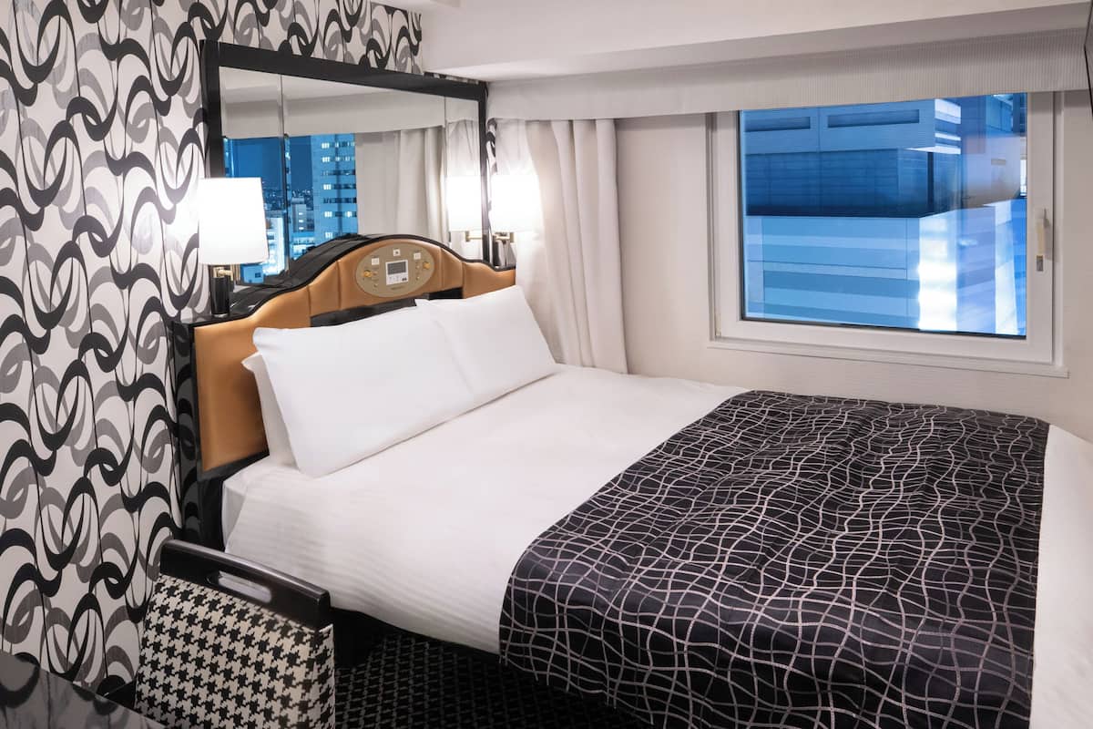 Une chambre d’hôtel avec un lit, une table de chevet, un miroir, une fenêtre donnant sur des immeubles et des rideaux à motifs.