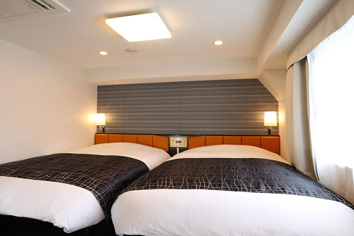 Une chambre d’hôtel avec deux lits, une tête de lit dotée d’un panneau de commande, des lampes de chevet et une fenêtre avec des rideaux.