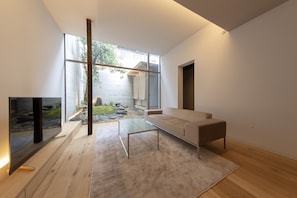 Een moderne woonkamer met een bank, een glazen salontafel en een televisie. Een groot raam biedt uitzicht op een tuin en een gebouw.
