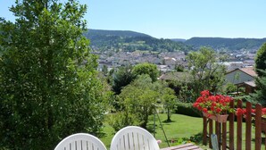 A patio with two white chairs, a wooden table, and a view of a town with houses and greenery in the background. - Charming 4* chalet with panoramic view, south-facing, sleeps 6. (Gérardmer)