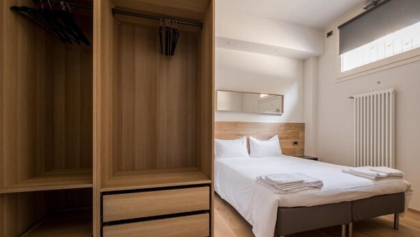 Une chambre avec une armoire en bois, un lit recouvert de draps blancs et un radiateur fixé au mur.