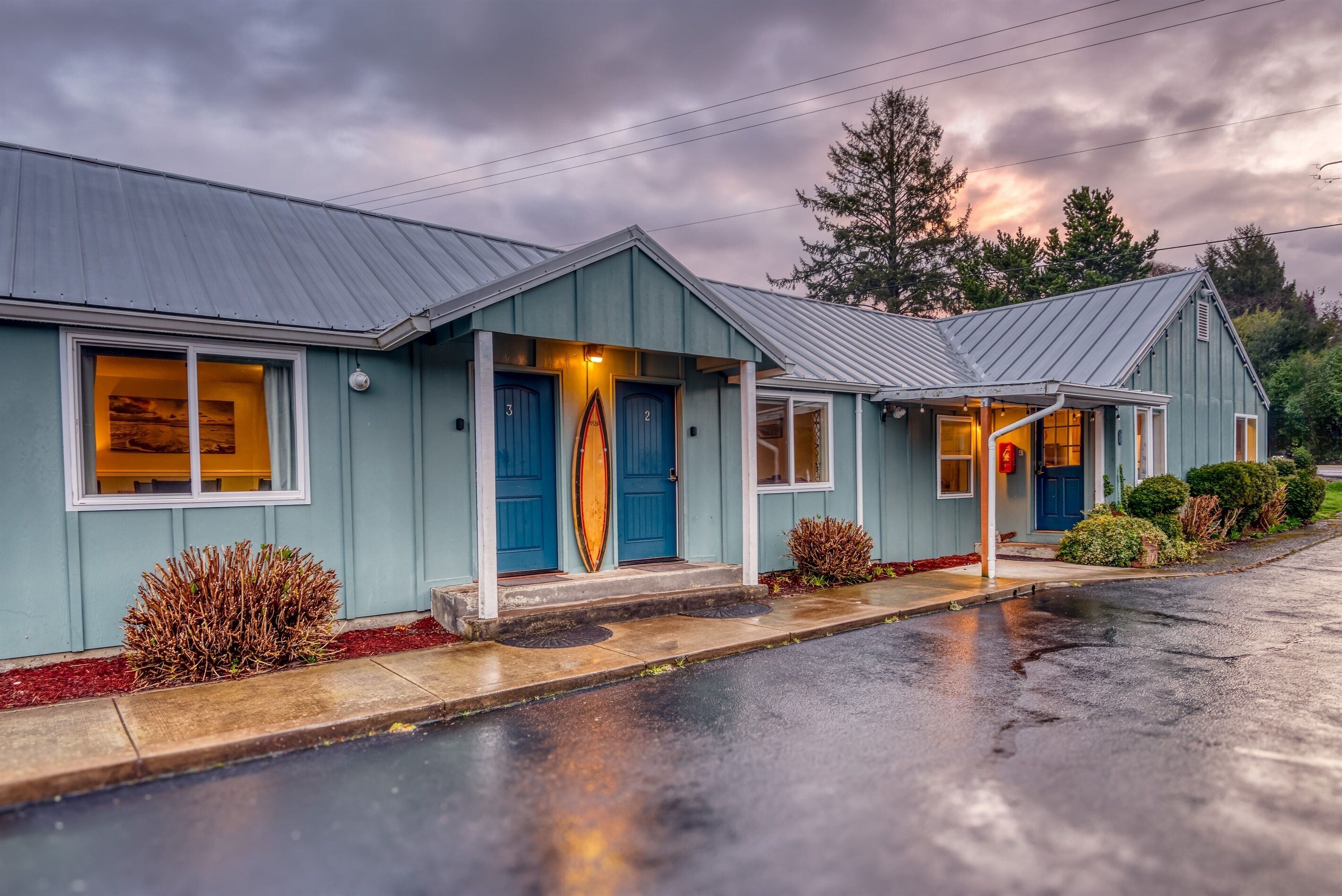 Une rangée de maisons bleues avec des portes numérotées, une planche de surf et une rue mouillée.