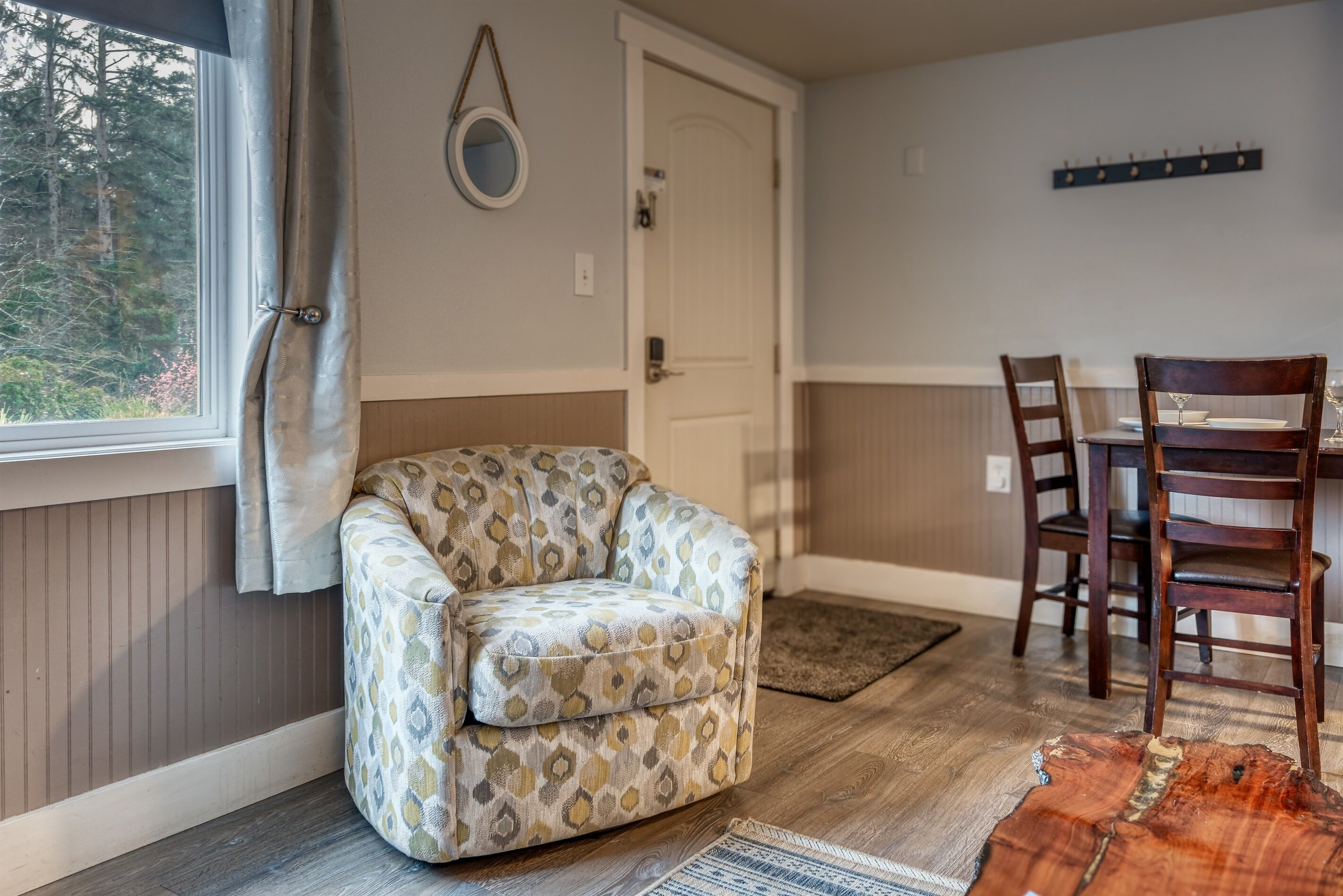 Un rincón acogedor con un sillón estampado, una mesa de comedor con sillas, suelo de madera, una puerta y una ventana con cortinas.