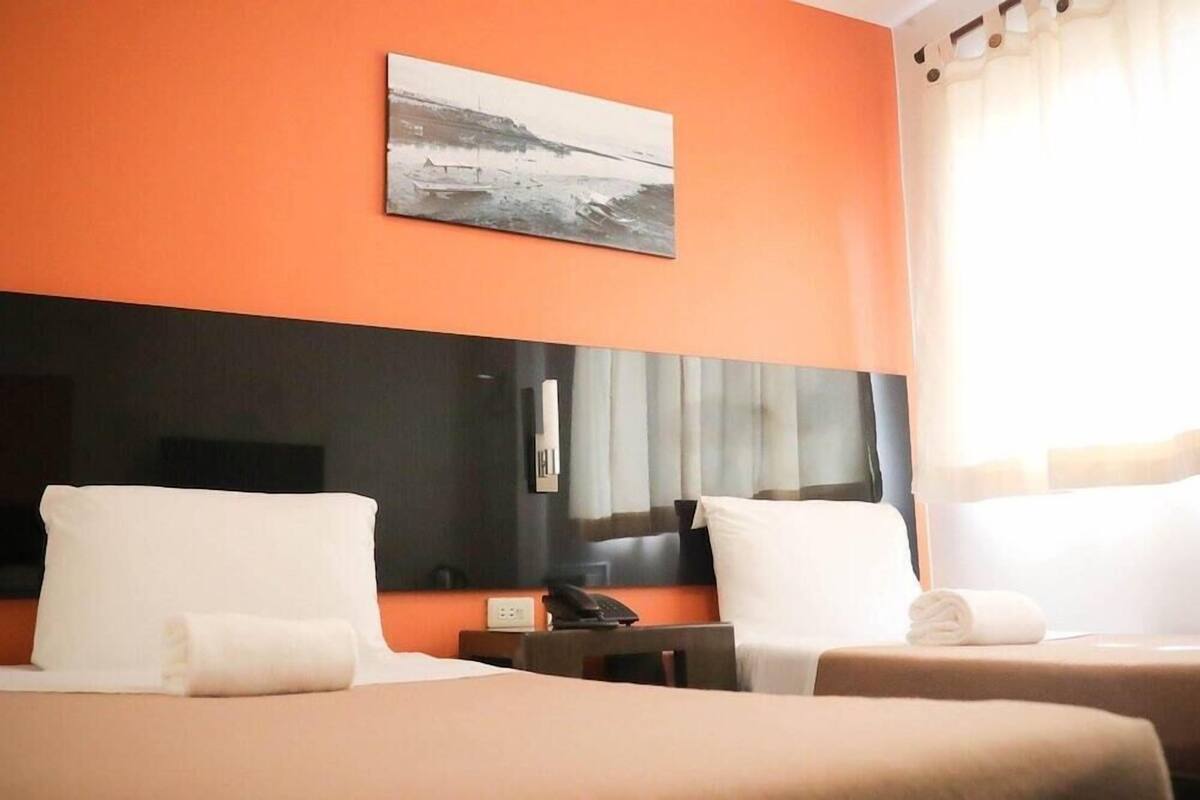 A hotel room with two beds, a large flat-screen TV, a painting on the wall, and a window with curtains.