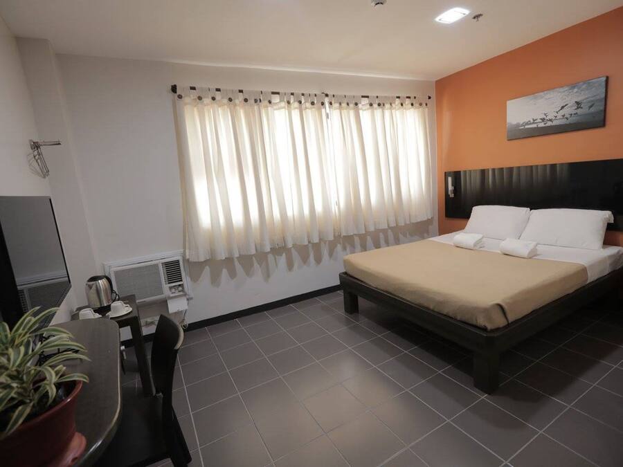 Une chambre d’hôtel avec un lit, un bureau et une chaise, une plante en pot, un téléviseur et une fenêtre avec des rideaux.