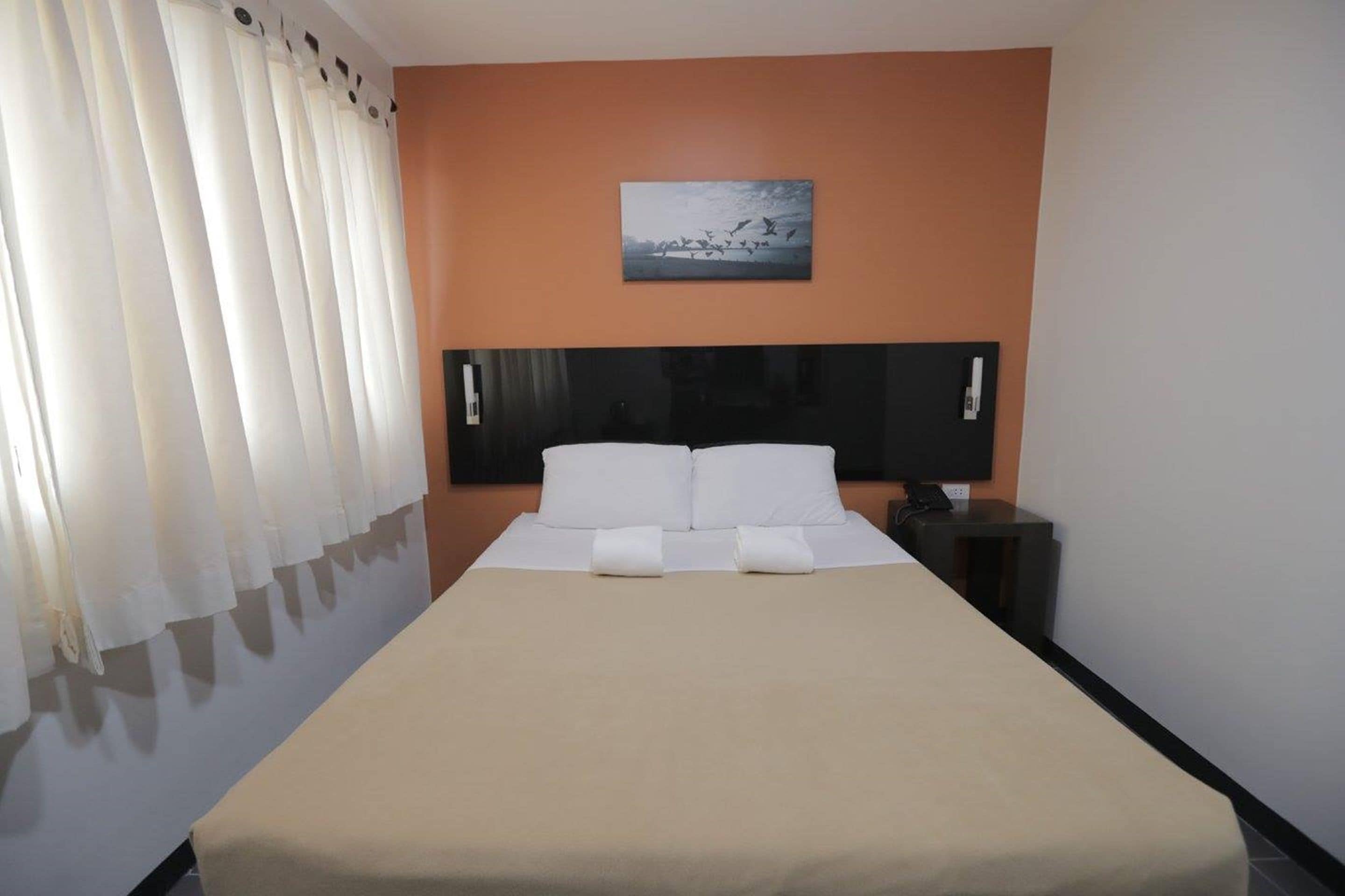 A hotel room with a bed, white curtains, a painting on the wall, and a nightstand.