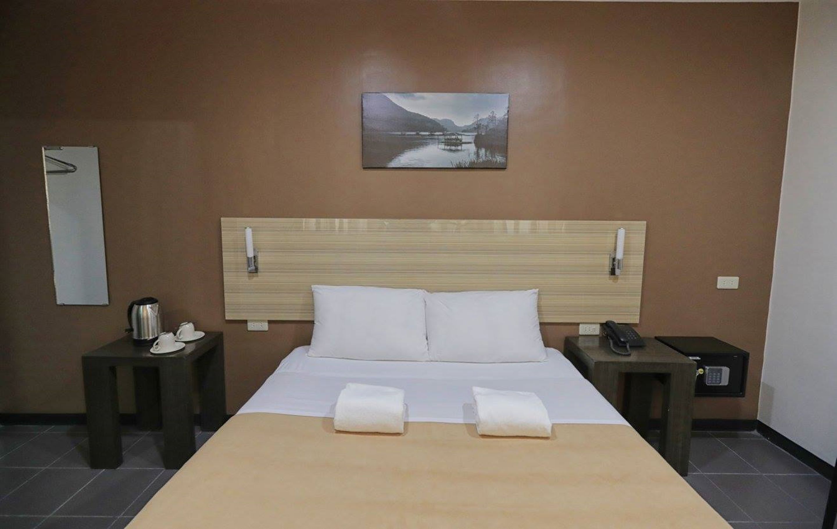 A hotel room with a bed, a painting of a landscape, a bedside table with a phone, and a small table with a kettle and cups.