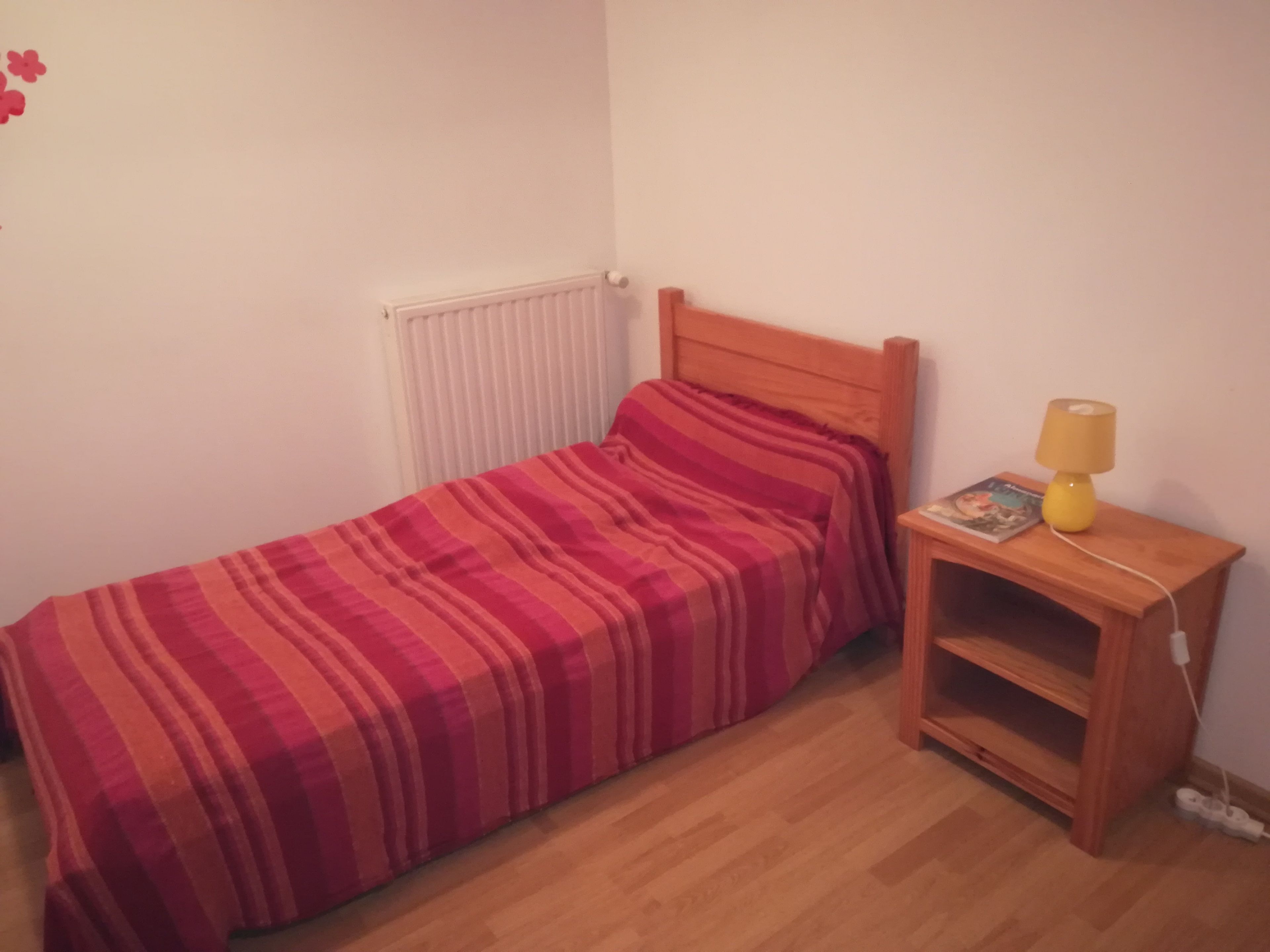 Une petite chambre avec un lit simple, une tête de lit en bois, une table de chevet avec une lampe et un radiateur.