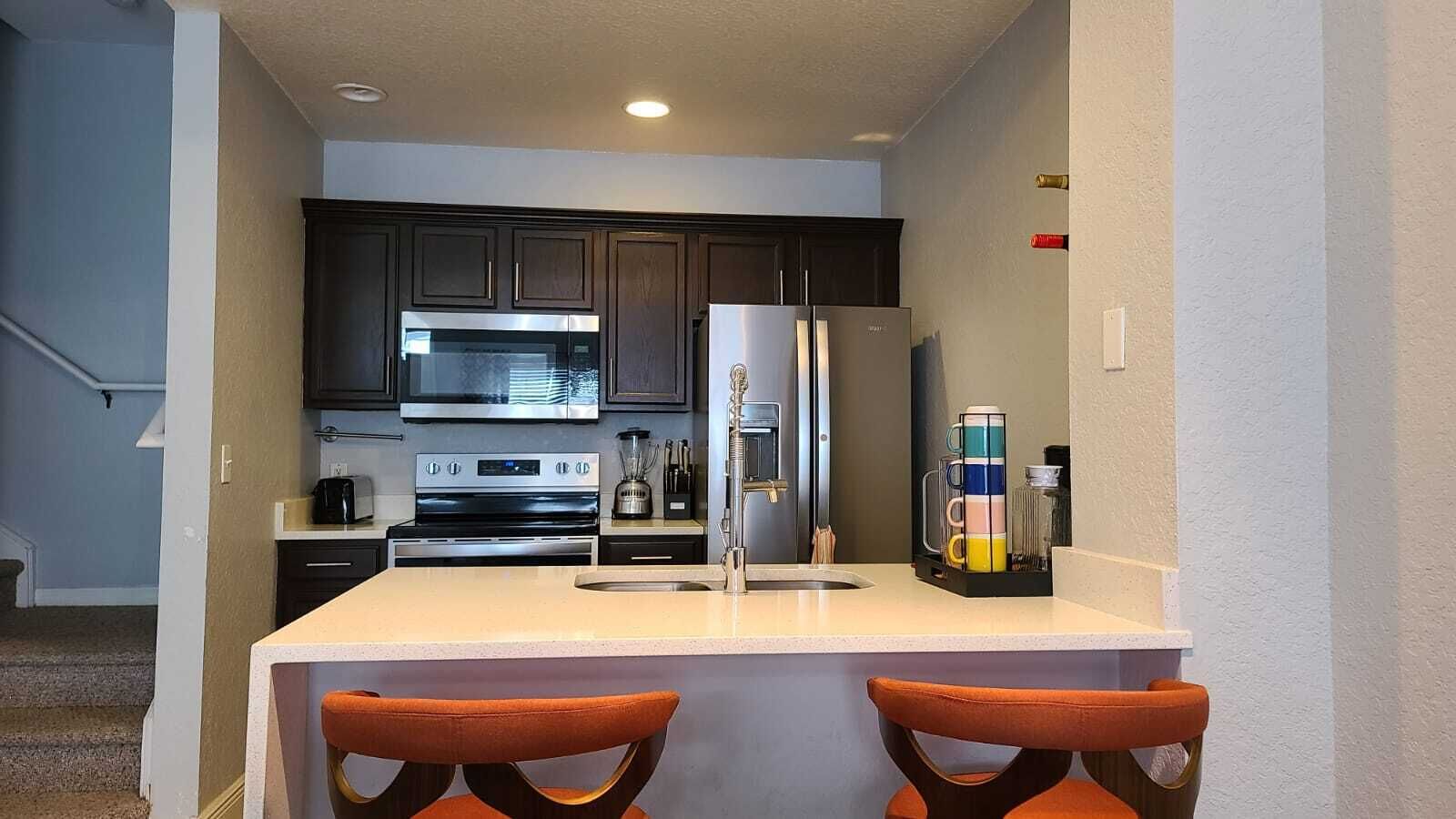 A compact kitchen with dark cabinets, stainless steel appliances, and a small island with two orange chairs.