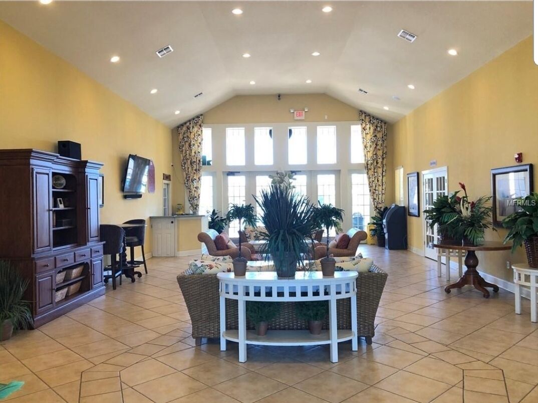 A spacious lobby with a high ceiling, large windows, a central round table with chairs, and potted plants.