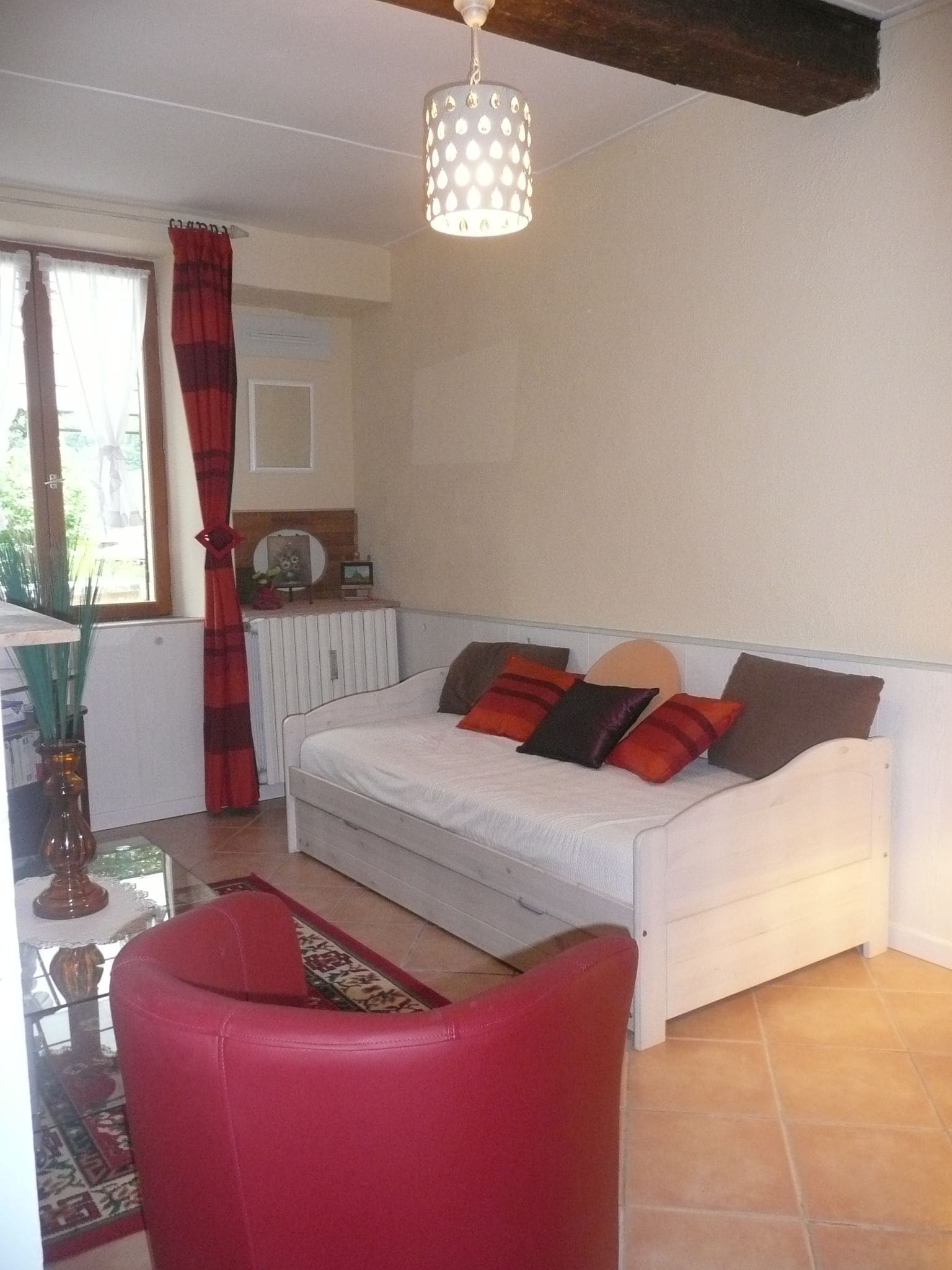 A bedroom with a white sofa bed, a red chair, a hanging light fixture, and a window with curtains.